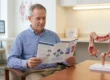 A medium shot photograph taken indoors with natural light coming from a large background window. In the center, a smiling middle-aged man with short grey hair, wearing a blue pinstriped button-down shirt, sits at a wooden table, holding and reading a folded medical education brochure. The brochure clearly displays titles "Colon Cancer Screening" and "Choosing Your Options" (text partially visible). To the right on the same table, a highly detailed, three-dimensional model of a human colon with a visible abnormality is positioned on a white plastic stand. The background is a clean medical office environment, with a desk, chair, a computer monitor displaying graphs (text illegible), and framed medical anatomical posters on the wall.