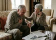 A medium eye-level shot captures a senior couple in a living room illuminated by soft, diffused daylight streaming from a window in the background. A man in a textured grey knit sweater sits on a vintage floral-patterned sofa, his face contorted in distress as he holds his chest. Beside him, a woman in a beige cardigan leans in with an urgent expression, holding a sleek black smartphone to her ear. The composition is balanced by a wooden coffee table in the foreground featuring a glass of water and folded newspapers with blurred headlines. On the far left, a square digital clock provides a sharp focal point, displaying the time as "15:05" in bold black digits.