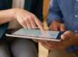 Close-up of two people's hands looking at a tablet. The person in the green blazer points to the screen, which is held by the person in the blue shirt. On the tablet screen, data visualizations like charts and graphs are clearly visible.