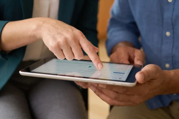 Close-up of two people's hands looking at a tablet. The person in the green blazer points to the screen, which is held by the person in the blue shirt. On the tablet screen, data visualizations like charts and graphs are clearly visible.