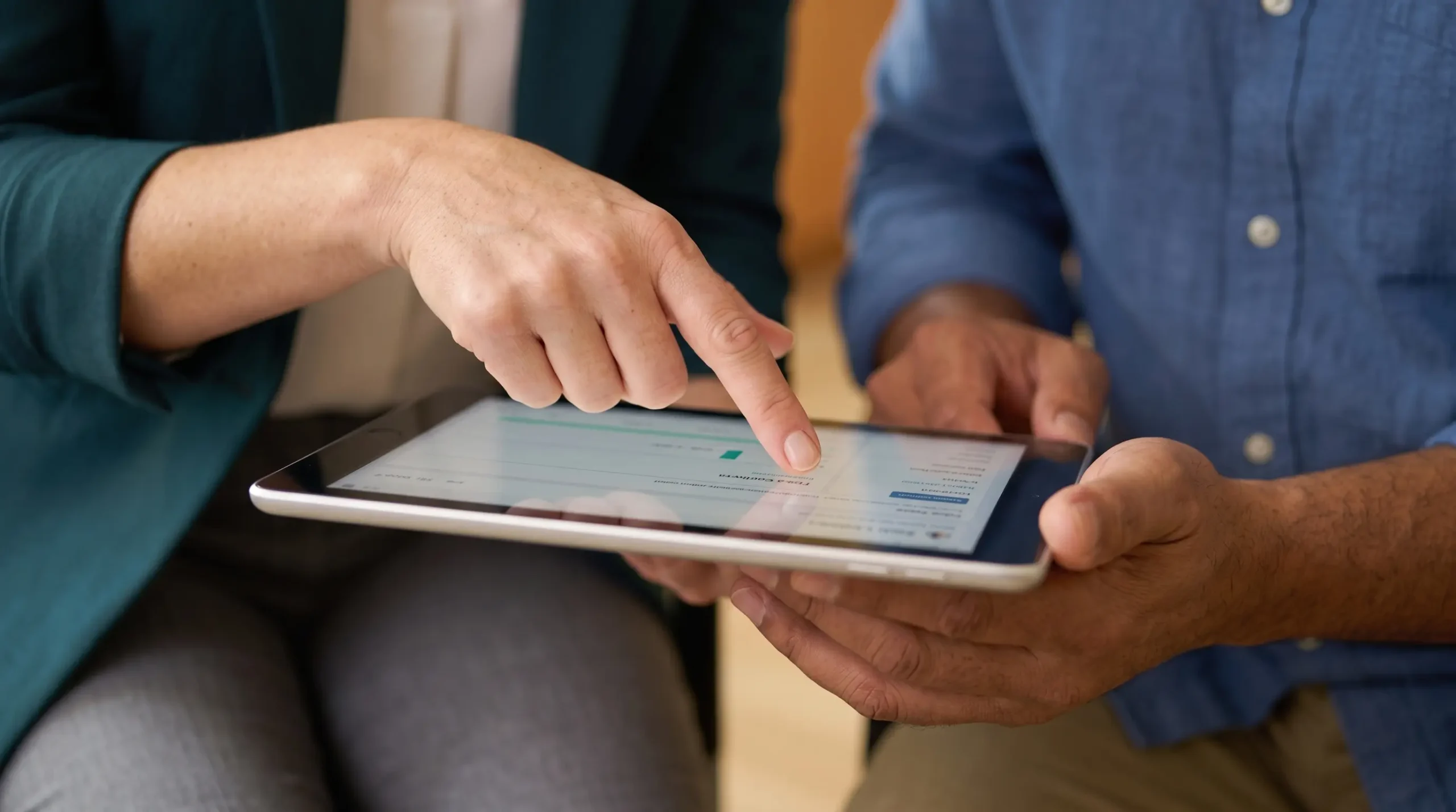 Close-up of two people's hands looking at a tablet. The person in the green blazer points to the screen, which is held by the person in the blue shirt. On the tablet screen, data visualizations like charts and graphs are clearly visible.