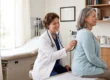 A bright, eye-level composition illuminated by soft natural sunlight streaming from a window on the left, casting a warm, airy, and inviting tone over a smiling female doctor examining a seated patient's back with a stethoscope. The image features a shallow depth of field that gently blurs the background exam table and wooden cabinets, emphasizing vibrant, true-to-life colors and a photorealistic aesthetic. The only text present is a tiny, indistinct embroidered logo or name on the doctor's white coat pocket.