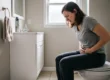 A medium shot in a softly lit domestic bathroom. Diffused natural daylight filters through a large window (or frosted pane) directly behind a woman, backlighting her and casting her distressed expression in soft detail. She is seated on a white toilet, leaning forward with a painful expression on her face and both hands pressed firmly against her lower abdomen. She wears a grey t-shirt and dark jogger pants. The frame is balanced with a white vanity unit and mirror to the left, holding a hand towel on a ring and a small bottle, over a tiled floor. The walls are a muted, neutral color. No visible text appears on any object.
